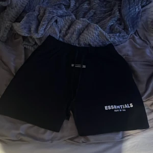 Svarta Essentials Fear of God shorts - Svarta shorts från Essentials Fear of God med vit logga tryckt på vänster ben. Modellen har elastisk midja med snörning och är tillverkad i ett mjukt material som känns skönt mot huden. Perfekta för chill stil.