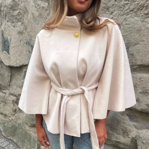 Trendig missäy poncho - Intressekoll på stilren beige kappa med knytbälte i midjan och vida, trekvartsärmar. Jackan har hög krage och en dekorativ guldfärgad knapp upptill. Perfekt för dig som gillar en clean och elegant look.