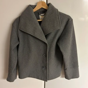 Grå kappa från Gina Tricot - Snygg grå kappa från Gina Tricot med bred krage och två stora knappar framtill. Jackan har en rak och lite boxig passform, perfekt för dig som gillar stilren och enkel design. Materialet känns mjukt och värmande, perfekt för kyligare dagar. Inte fri frakt! 