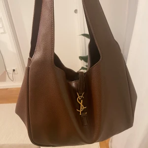 Brun axelväska - Säljer en brun axelväska i lyxigt skinn med guldigt YSL-spänne framtill. Väskan har bred axelrem, rymligt huvudfack och innerficka med dragkedja. Perfekt för dig som vill ha en stilren och klassisk väska med exklusiv känsla.