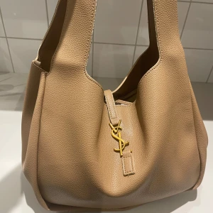Beige axelväska  - Säljer en snygg beige axelväska från Saint Laurent i mjukt lyxigt skinn. Väskan har breda axelband och ett YSL-spänne i guld framtill. Perfekt storlek för att få plats med allt du behöver och ger en clean och lyxig vibe.