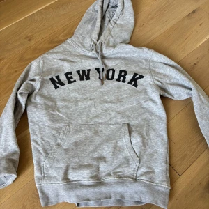 Grå hoodie med New York-tryck - Snygg grå hoodie med svart 'NEW YORK'-text över bröstet. Tröjan har huva med snörning, magficka och ribbade muddar. Perfekt för chill dagar eller när du vill ha en avslappnad streetstyle-look.