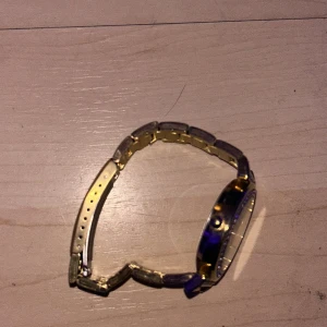 Guldig klocka med stenar från Regal - Snygg guldig klocka från Regal med metallarmband och glittrande stenar runt urtavlan. Urtavlan är ljus med guldiga visare och index. Klassisk rund form och stilren design som drar blickarna till sig.