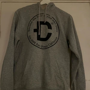 Grå hoodie med tryck San Diego - Snygg grå hoodie med stort svart tryck framtill där det står 'Become the hero of your own story' och 'San Diego California'. Tröjan har huva med snören och en klassisk känguruficka. Perfekt för chill dagar eller när du vill ha en avslappnad stil.