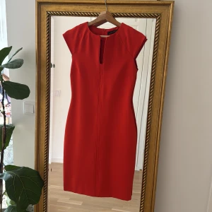 Orange röd kortklänning från Zara Woman - Snygg röd kortklänning från Zara Woman med stilren design och markerade axlar. Klänningen är ärmlös, har en liten slits vid halsringningen och dragkedja bak. Perfekt för dig som vill sticka ut med en enkel men elegant look.