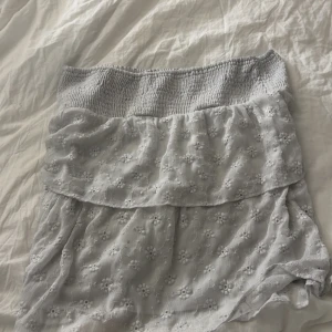 Ljusgrå blommig volangkjol - Supersöt ljusgrå kjol med broderade blommor och volangdetaljer. Kjolen har en smockad midja som sitter bekvämt och ger en fin passform. 