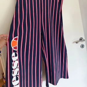 Randig byxdress från Ellesse, marinblå - Säljer en snygg byxdress från Ellesse i marinblått med röda och vita ränder. Byxdressen har breda ben, v-ringning och axelband. Stor logga på ena benet. Perfekt för dig som gillar sportig och trendig stil.