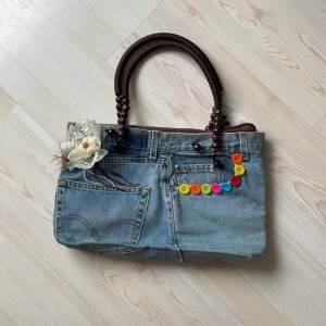 Jeans handväska med färgglada knappar - Unik handväska i jeans från Charkorn med bruna handtag i träpärlor, dekorerad med färgglada knappar och en spetsblomma. Väskan har flera fickor och dragkedja upptill. Perfekt för dig som gillar kreativ och personlig stil.