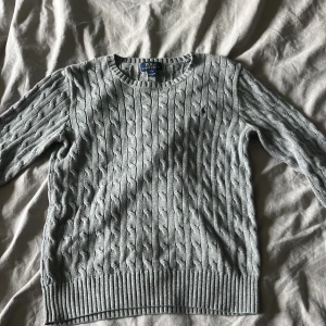 Grå kabelstickad tröja Polo Ralph Lauren - Kabelstickad grå tröja från Polo Ralph Lauren med rund halsringning och broderad logga på bröstet. Tröjan har långa ärmar och ribbade muddar vid ärmslut. Pris är inte hugget i sten, var inte rädd att lägga ett prisförslag.