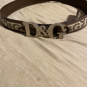 Brunt D&G bälte med strassspänne - Snyggt brunt bälte från Dolce & Gabbana med stort D&G-spänne i silver dekorerat med strass. Bältet har logomönster längs hela bandet och är gjort i skinn med kontrastsömmar. Perfekt för att lyfta din outfit med en lyxig touch.