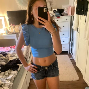 Blå croppad topp med volangärmar från H&M - Supersöt ljusblå croppad topp från H&M med volangdetaljer på axlarna. Toppen är ärmlös och har en tight passform som framhäver midjan. Perfekt för dig som vill ha en trendig och lekfull look i sommar.