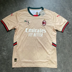 AC Milan Bortatröja – Emirates Edition (Puma) - AC Milan bortatröja i exklusiv beige/guld nyans med detaljer i rött och grönt. Säsongens officiella tröja från Puma med “Fly Better”-spons. Original med tags kvar! 🧼 Helt ny med taggar 📏 Storlek: 1M Regular ⚽️ För fans, samlare eller stilmedvetna