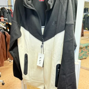 Nike svartvit TECH DRESS med dragkedja - Har alla storlekar!  Nike dress i svart och vitt med snygg färgblock-design. Jackan har hel dragkedja framtill, två fickor med dragkedja och huva. Tillverkad i bomull och polyester för en skön känsla. Perfekt för dig som gillar sportig och stilren look.