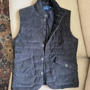 Ralph Lauren vest - Snygg svart quiltad dunväst från Polo Ralph Lauren med ståkrage och stora fickor framtill. Västen har knäppning med knappar och är tillverkad i mocka. Perfekt för lager-på-lager och stilren look. Köpt på rea för 8,300kr. Storlek S men anser att den passar M
