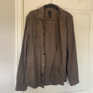 Rutig brun skjorta från H&M - Snygg rutig skjorta från H&M i bruna och beige toner med inslag av blått och svart. Skjortan har klassisk krage, bröstficka och knäppning framtill. Regular fit och långärmad – perfekt för en chill och stilren look.