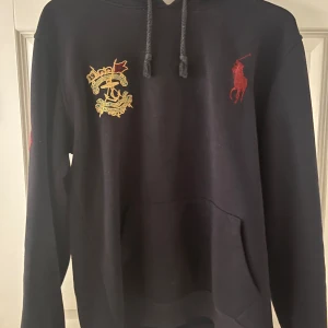 Svart hoodie från Polo Ralph Lauren - Svart hoodie från Polo Ralph Lauren med broderad guldfärgad logga och röd polospelare på bröstet. Stor röd siffra 3 på ärmen. Klassisk känguruficka och huva med snörning. Perfekt för dig som gillar sportig och stilren streetwear.