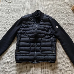 Svart Moncler pufferjacka med dragkedja - Säljer en svart pufferjacka från Moncler med quiltad framsida och släta ärmar. Jackan har två dragkedjefickor, hög krage och hel dragkedja framtill. Perfekt för höst och vinter, riktigt snygg och stilren design med Moncler-logga på ärmen.