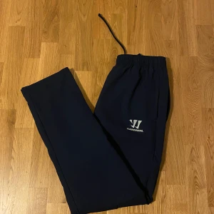 Mörkblå täckbyxor från Warrior - Mörkblå täcksbyxor från Warrior med elastisk midja och snörning. Byxorna har en enkel och clean look med Warrior-logga på ena benet. Perfekta för utomhusaktiviteter, träning eller kallare dagar, tillverkade i mjukt och bekvämt material.