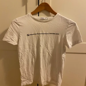 Vit Calvin Klein Jeans t-shirt - Vit t-shirt från Calvin Klein Jeans med svart minimalistiskt tryck och logga på bröstet. Klassisk rund halsringning och korta ärmar. Tillverkad i mjuk bomull för en clean och enkel look.