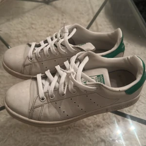 Adidas Stan Smith vita sneakers - Klassiska Adidas Stan Smith sneakers i vitt skinn med gröna detaljer på hälen och tungan. Skorna har rund tå, platt sula och snörning framtill. Perfekta för en clean och tidlös look, passar till det mesta i garderoben.