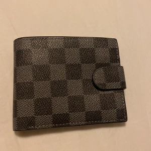 Rutig svart plånbok i skinn - Snygg svart plånbok med rutigt mönster och praktisk knappstängning. Plånboken har flera kortfack och ett fönster för ID eller körkort. Tillverkad i skinn och har en stilren, modern look som passar perfekt i fickan.