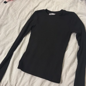 Svart ribbad långärmad tröja F.A.Sister - En svart, ribbad långärmad tröja från F.A.Sister. Tröjan har en rund halsringning och är figurnära i modellen. Perfekt basplagg med material som passar till allt. Enkel och stilren design som funkar året runt.