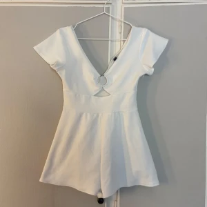 Vit kort shortsdress från H&M Divided - Supersöt vit kort shortsdress från H&M Divided med v-ringning och snygg ringdetalj framtill. Klänningen har korta ärmar och en lätt utsvängd kjol. Perfekt för sommarens alla tillfällen och ger en fräsch och trendig look.