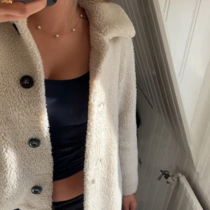 Beige teddyjacka med stora knappar - Supermysig beige teddyjacka med stora svarta knappar framtill. Jackan har en klassisk krage och är tillverkad i fluffigt teddy-material som håller dig varm. Perfekt för dig som vill ha en chill och trendig look under höst och vinter.