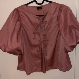 Rosa blus med puffärmar och knyt - Supersöt rosa blus från ginatricot med puffiga ärmar och två knyt framtill. Blusen har en lätt glansig finish och en luftig passform. Perfekt för dig som gillar statement-ärmar och unika detaljer.