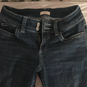 Mörkblå bootcut jeans från Nelly - Snygga mörkblå jeans från Nelly med bootcut passform och låg midja. Jeansen har vita kontrastsömmar, dubbla knappar i midjan och coola bakfickor med vit zigzag-detalj. Perfekta för dig som gillar en klassisk men trendig look. De är stretchiga i materialet och är väldigt bekväma att ha på sig. Har använt de några enstaka gånger men inget har slitits.