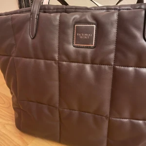 Quiltad brun handväska Victoria's Secret - Stor quiltad handväska från Victoria's Secret i mörkbrunt skinnliknande material. Väskan har ljusrosa foder och handtag med både brun och ljus sida. Perfekt för dig som vill ha en rymlig och snygg väska med exklusiv känsla. Insidan är relativt smutsig som jag inte lyckats ta bort. Därav blir det en prissänkning. Annars finns det inga defekter. 