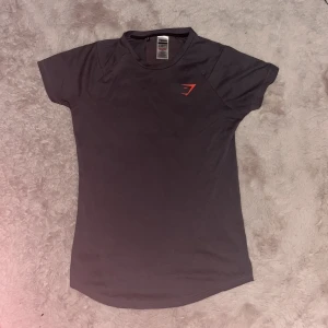 Svart Gymshark t-shirt med röd logga - Svart t-shirt från Gymshark i sportig modell med korta ärmar. Stor röd Gymshark-logga på ryggen och mindre logga på bröstet. Tillverkad i mjuk polyester, perfekt för träning och aktiva dagar.