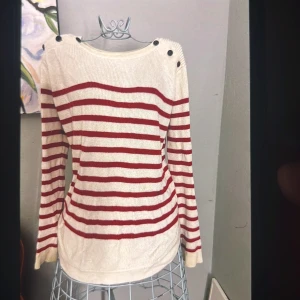 Randig stickad tröja med knappar - Vit och röd randig stickad tröja med långa ärmar och dekorativa svarta knappar på axlarna. Tröjan har en rund halsringning och är mjuk skön