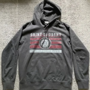 Svart Saint Laurent hoodie med tryck - Säljer nu denna gråa hoodie från Saint Laurent i mycket fint skick nästintill som ny. Säljer då den blivit för liten storlek s men passar dig som har xs. Hör av er vid frågor eller funderingar priset går att diskutera!!