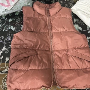 Rosa dunväst med dragkedja - Säljer en puffig rosa dunväst med hög krage och dragkedja framtill. Västen har två stora fickor på sidorna och är quiltad för extra volym. Perfekt att slänga över en hoodie för en chill look.