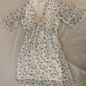 Blommig volangklänning från Pigalle  - Supersöt kort blommig klänning från Pigalle i vitt med små rosa och blå blommor. Klänningen har v-ringning med volangdetaljer, korta volangärmar och markerad midja. 