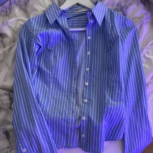 Blåvit randig skjorta från Bershka - Snygg blå skjorta med vita ränder från Bershka. Skjortan har klassisk krage, lång ärm och knappar framtill. Figurnära passform som ger en clean och fräsch look. Perfekt till jeans eller kjol för en trendig stil.