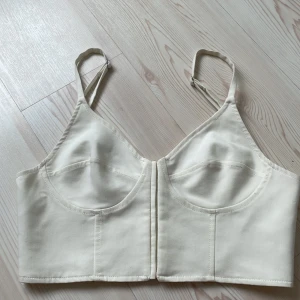 Vit korsett-topp från Zara - Snygg vit korsett-topp från Zara med justerbara axelband och hakknäppning framtill. Tjockare bomullsmaterial med markerade sömmar och formade kupor för en clean och stilren look. Perfekt att styla med jeans eller kjol för en trendig vibe.