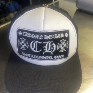 Chrome Hearts ”CH” truckerkeps svart/vit - Snygg truckerkeps från Chrome Hearts i svart och vitt med stor broderad patch framtill. Kepsen har mesh-bakstycke och böjd skärm. Märkesdetaljer på insidan och klassisk streetwear-vibe. Perfekt för dig som gillar statement-plagg och exklusiva accessoarer.