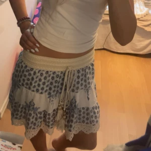 Snygg mönstrad kjol - Supersöt kortkjol i vitt och blått med bohemiskt mönster och broderade detaljer. Kjolen har bred midja med snörning och tofsar, samt volangkant nertill. Perfekt för sommarens alla äventyr! Köpt på en marknad i Kroatien så har tyvärr inget märke men ändats använd en gång💕💕