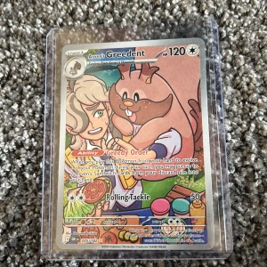 Arven's Greedent - Cool Pokémonkort med färgstark illustration av Arven och Greedent. Perfekt för samlare och fans av Pokémon! Kortet har specialeffekter och är skyddat i plastfodral. Ett måste för dig som vill ha något unikt i din samling.
