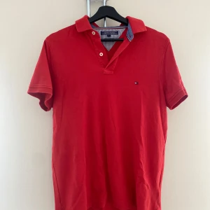 Röd Tommy Hilfiger Piké  - Röd piké t-shirt från Tommy Hilfiger i slim fit-modell. Klassisk krage med två knappar och diskret broderad logga på bröstet. Tillverkad i bomull med pikéstruktur och korta ärmar. Snygg och stilren för en clean look.