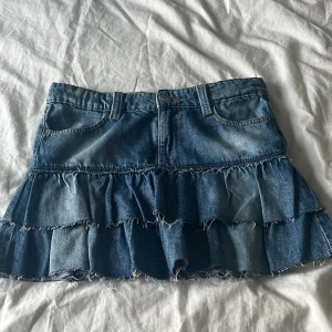 Jeanskjol med volanger från Gina Tricot - Supersöt jeanskjol från Gina Tricot med låg midja och volanger i två lager. Kjolen är blå, har klassiska jeansfickor och råa kanter för en cool vibe. Perfekt för dig som gillar denim och vill ha en trendig look. Aldrig använd + lapp kvar. Storlek 158 och har justerbar midja. Mått: 37 cm rakt över midjan, 32 cm lång fram. 💗