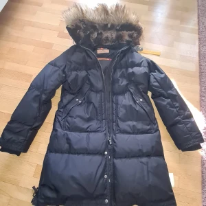 Svart lång dunjacka med fuskpäls - Säljer en svart lång dunjacka från Parajumpers med snygg fuskpäls på huvan. Jackan har dragkedja, knappar och två stora fickor framtill. Perfekt för kalla vinterdagar och har en klassisk, stilren look.