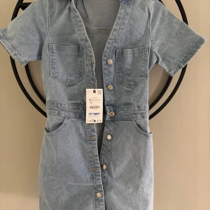 Ljusblå jeansklänning med knappar - Snygg ljusblå jeansklänning från Zara i kort modell. Klänningen har korta ärmar, fyra fickor och knappar hela vägen framtill. Perfekt för dig som gillar en avslappnad men trendig look.