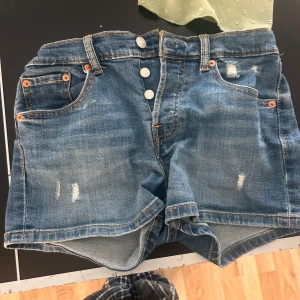 Levi's blå jeansshorts med knappar - Klassiska blå jeansshorts från Levi's med slitna detaljer och fyra synliga knappar framtill. Shortsen har fem fickor, bälteshällor och den ikoniska Levi's-lappen bak. Perfekta för en avslappnad stil under varma dagar.