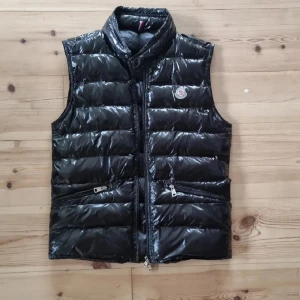 Svart dunväst från Moncler - Snygg svart dunväst från Moncler med glansig finish och klassisk logga på bröstet. Västen har hög krage, två dragkedjefickor och quiltad design. Perfekt att slänga över en hoodie eller tröja för extra värme.