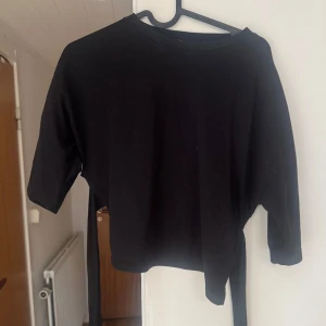 Svart croppad topp med knytband - En svart croppad topp med trekvartsärmar och snygga knytband i midjan som ger en cool omlottkänsla. Perfekt för att styla med höga jeans eller kjol. Enkel och stilren design som passar till många looks.