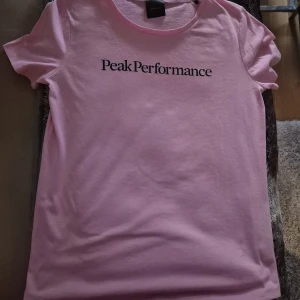 Rosa Peak Performance t-shirt - Säljer en rosa t-shirt från Peak Performance med svart logga på bröstet. Klassisk rund halsringning och korta ärmar. Skön och mjuk i bomull, perfekt till jeans eller shorts. Enkel och stilren design som funkar året runt.