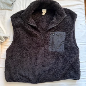Svart fleeceväst med bröstficka - Mysig svart fleeceväst med hög krage och en praktisk bröstficka med dragkedja. Västen har en half zip framtill och är perfekt för lager-på-lager-stil. Använd en gång 